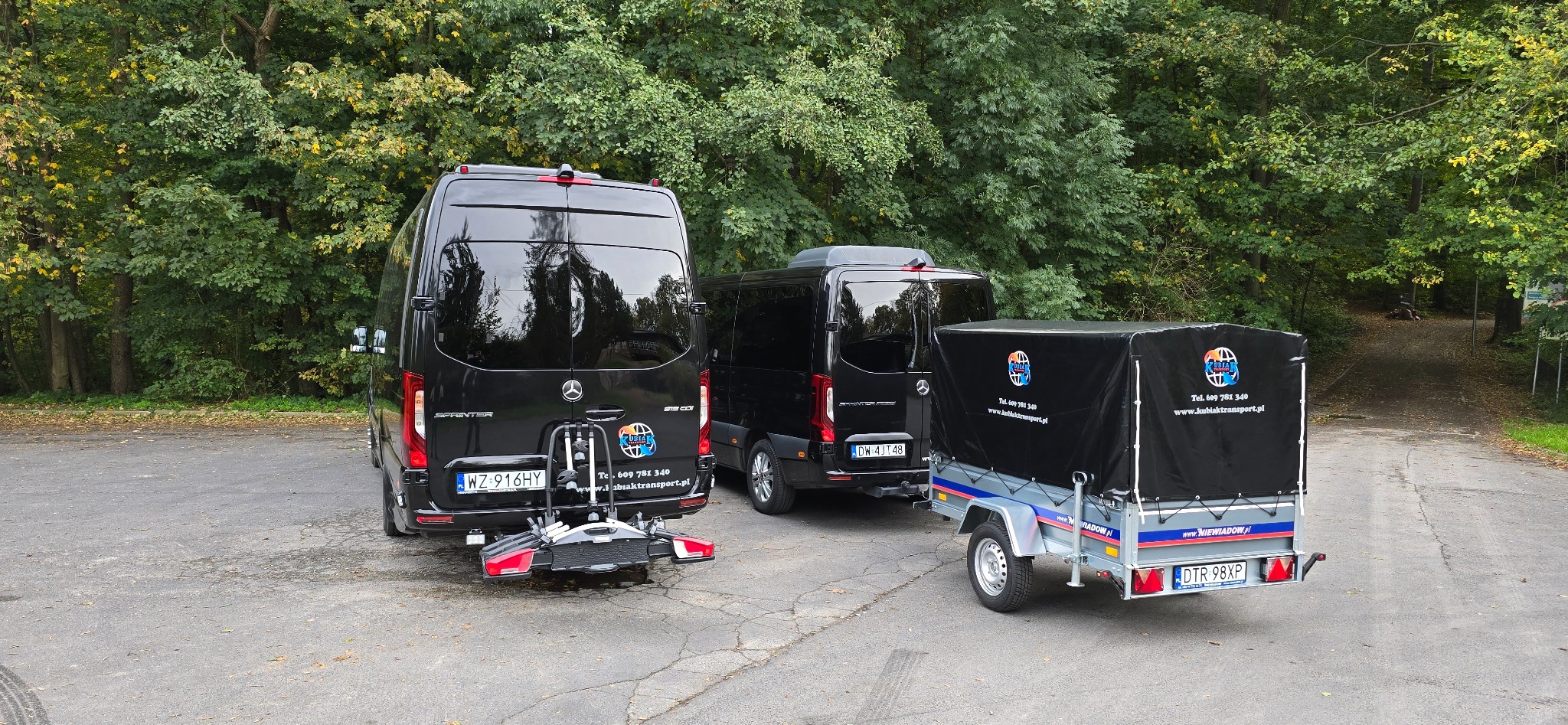 Dwa czarne busy Mercedes Sprinter oraz przyczepa z czarną plandeką na tle drzew, widok z tyłu, widoczne numery rejestracyjne i logo firmy transportowej.
