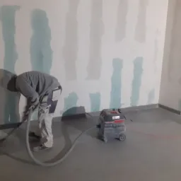 Pracownik w kombinezonie szlifuje ścianę po szpachlowaniu za pomocą szlifierki z odkurzaczem przemysłowym, widoczne pionowe pasy zagruntowanej ściany.