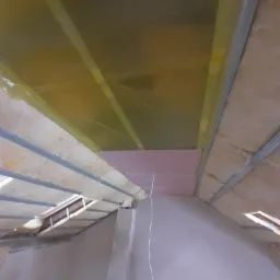 Poddasze w trakcie ocieplania: widoczna wełna mineralna, folia paroizolacyjna, metalowy stelaż i fragment różowej płyty termoizolacyjnej, ujęcie z dołu.