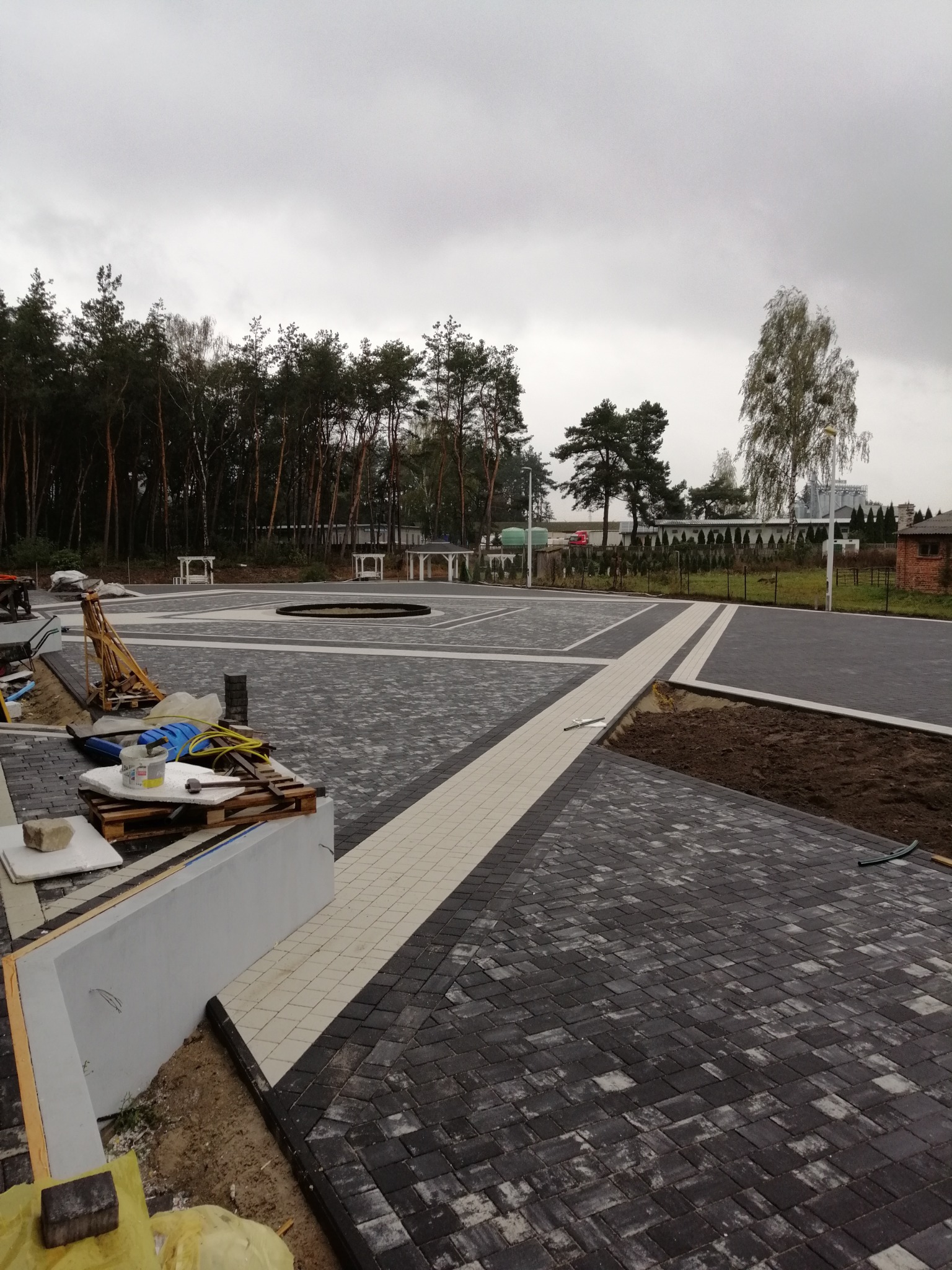 Rozległy plac z ułożonej kostki brukowej w odcieniach szarości i bieli, z elementami dekoracyjnymi w postaci okręgu i prostokątnych obszarów, widoczne narzędzia i materiały budowlane, w tle drzewa...
