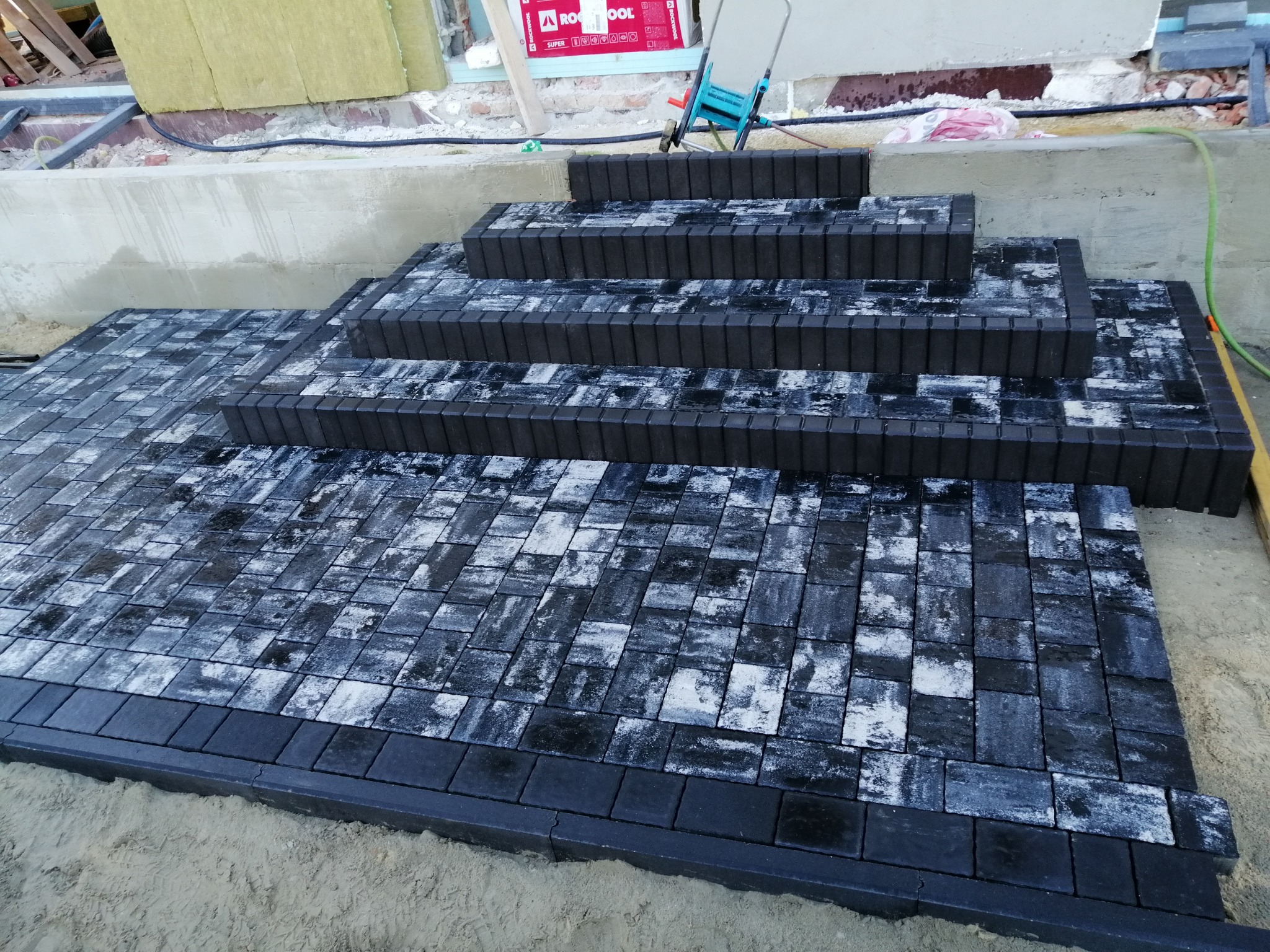 Wykończone schody zewnętrzne z czarno-białej kostki ułożonej na piasku, z widocznymi elementami konstrukcyjnymi budynku w tle.