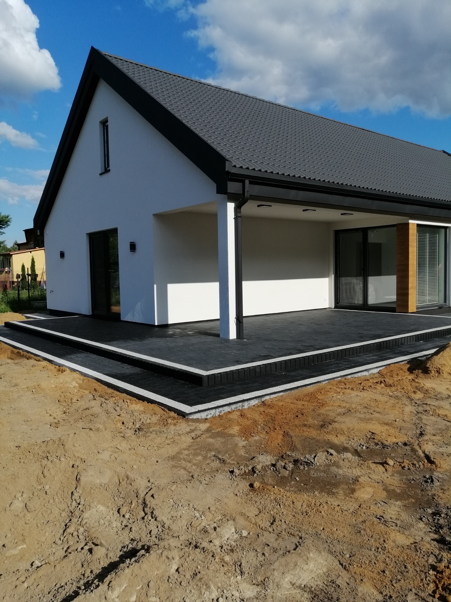 Nowoczesny dom z tarasem wyłożonym czarną kostką brukową z jasnoszarą obwódką, widoczne przygotowane podłoże z piasku.