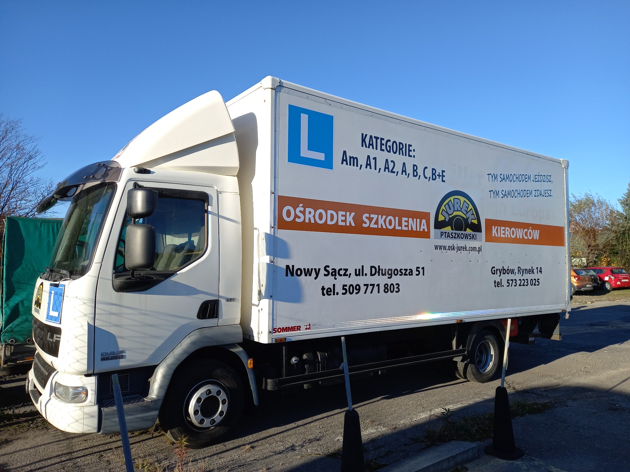 Biały samochód ciężarowy z oznaczeniami ośrodka szkolenia kierowców, widoczne kategorie prawa jazdy: Am, A1, A2, A, B, C, B+E, adres i telefon kontaktowy.