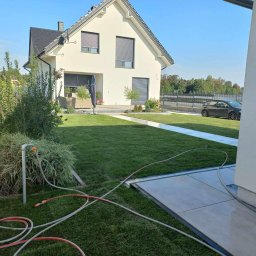 Mateusz Firaza - Nowoczesny dom z zadbanym trawnikiem i systemem nawadniania w trakcie pracy, widoczne węże ogrodowe. Trawa krótko skoszona, z tyłu zaparkowany samochód.
