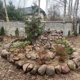 Mateusz Firaza - Ogród skalny z kaskadą ułożoną z kamieni różnej wielkości, otoczony obwódką z większych kamieni, z młodymi drzewkami iglastymi i liściastymi, na tle szarego, betonowego ogrodzenia i domu.