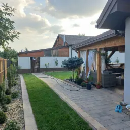 Ujęcie ogrodu z perspektywy ścieżki, ukazujące świeżo założony trawnik, ozdobne krzewy wzdłuż ogrodzenia, altanę z zasłonami i narzędzia ogrodnicze w tle.