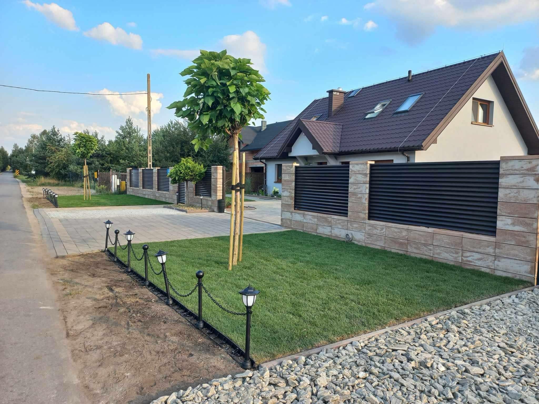 Nowoczesny ogród frontowy z równo przystrzyżonym trawnikiem, młodymi drzewami podpartymi palikami i ogrodzeniem z kamiennych bloczków oraz metalowych paneli, oświetlony ozdobnymi lampami łańcuchowymi.