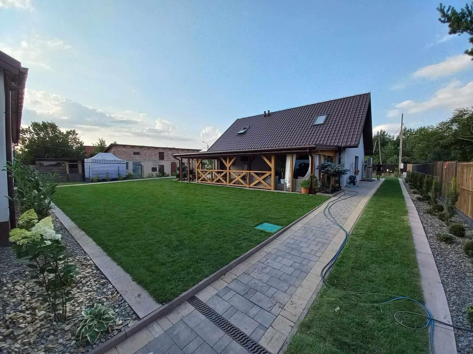 Rozległy, zadbany trawnik przed domem z drewnianą werandą, obrzeża z kamienia i kostki brukowej, widoczny budynek gospodarczy w tle.