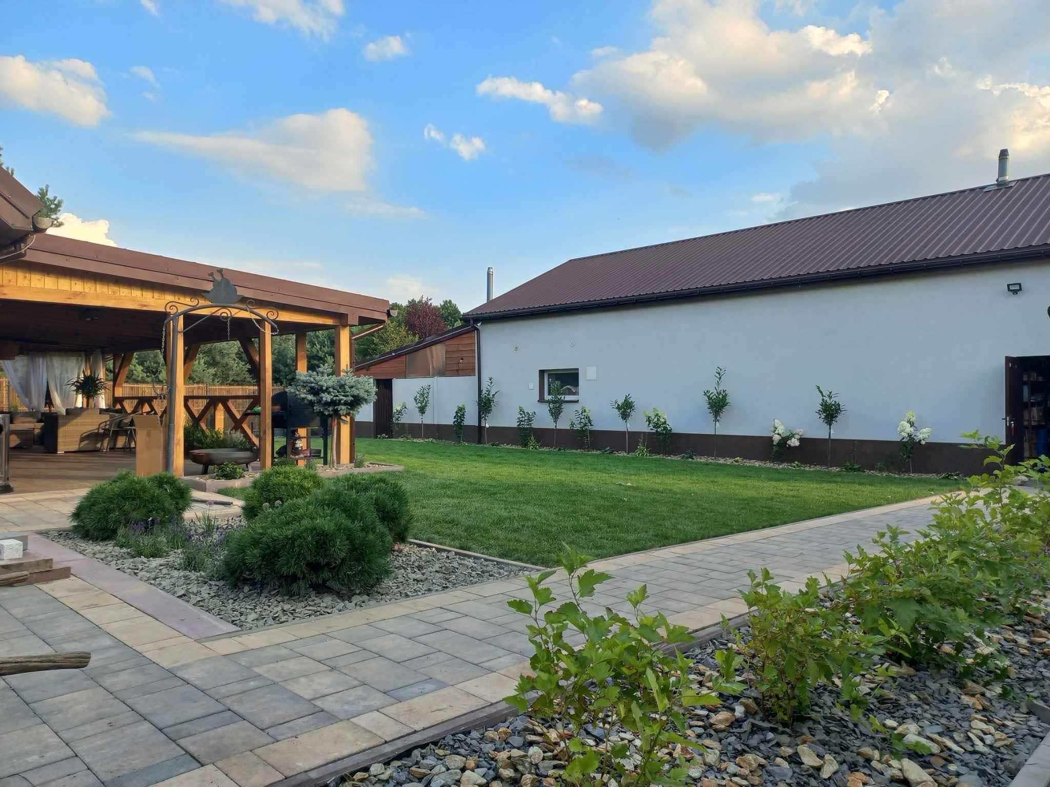Uporządkowany ogród z równo przystrzyżonym trawnikiem, ścieżką z kostki brukowej, ozdobnymi krzewami i altaną z drewnianą konstrukcją, na tle budynku z brązowym dachem i rzędem młodych drzewek...