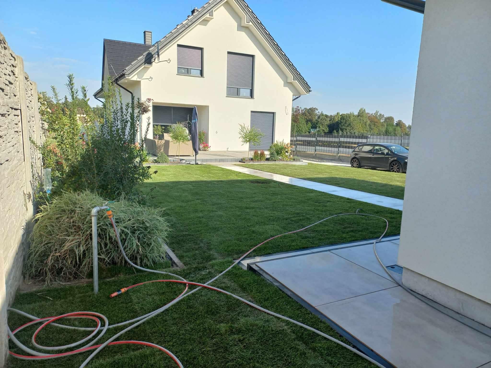 Nowoczesny dom z zadbanym trawnikiem i systemem nawadniania w trakcie pracy, widoczne węże ogrodowe. Trawa krótko skoszona, z tyłu zaparkowany samochód.