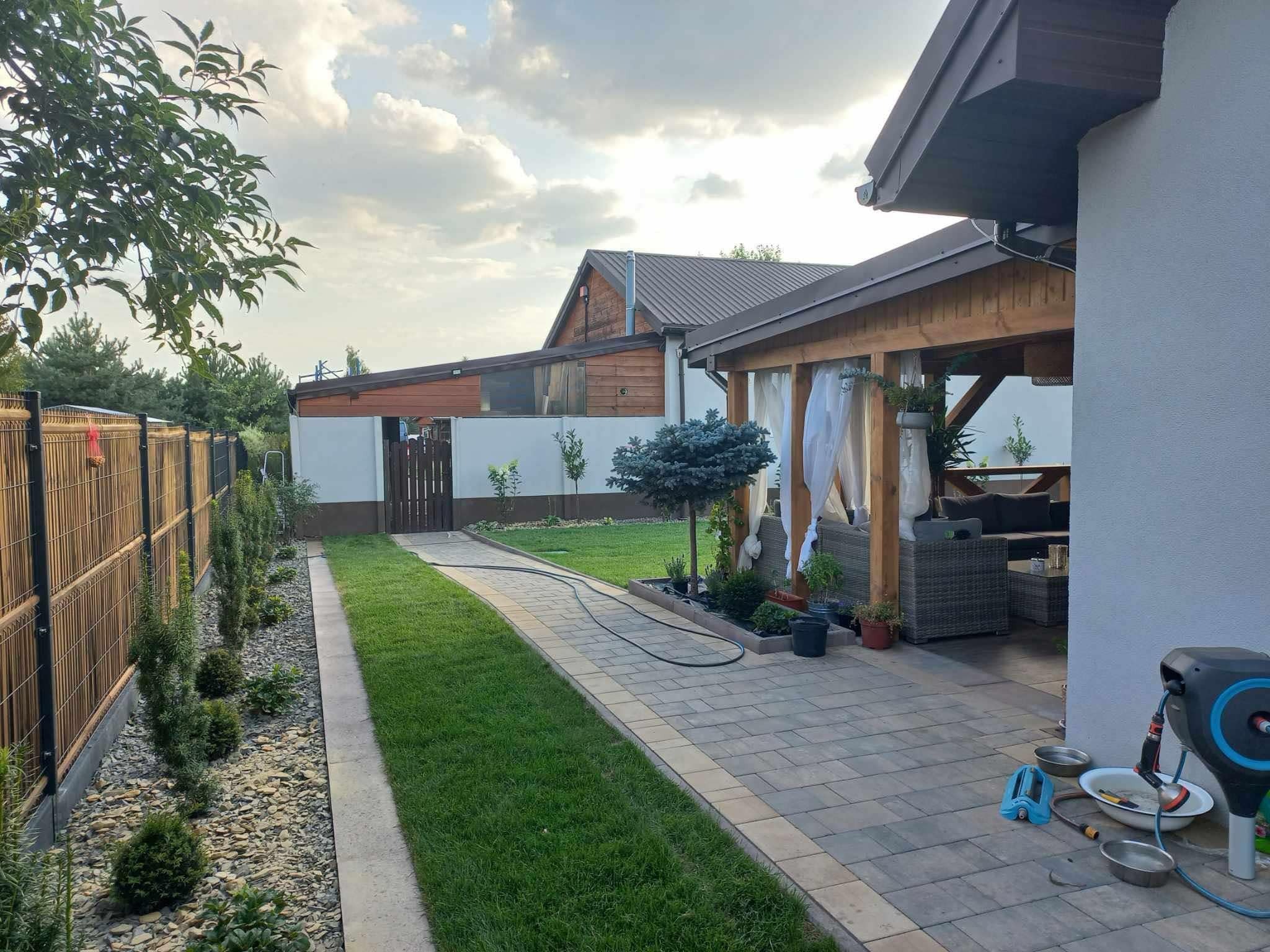 Ujęcie ogrodu z perspektywy ścieżki, ukazujące świeżo założony trawnik, ozdobne krzewy wzdłuż ogrodzenia, altanę z zasłonami i narzędzia ogrodnicze w tle.