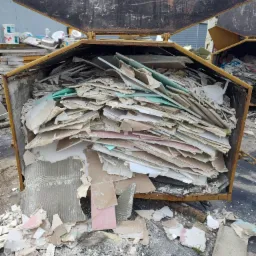 Żółty kontener wypełniony po brzegi gruzem budowlanym, głównie fragmentami płyt kartonowo-gipsowych z resztkami farb i tapet, widok z góry na otwarty kontener, na ziemi wokół porozrzucane mniejsze...