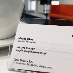 Wizytówka eksperta finansowego Magdy Okroj z Open Finance S.A., prezentowana w przezroczystym stojaku, z widocznym numerem telefonu i adresem e-mail, obok filiżanki i szklanki.