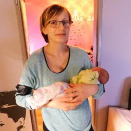 Kobieta w okularach trzyma noworodka w ramionach, w tle różowe ściany z dekoracjami motyli i napisami.