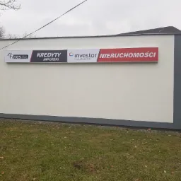 Zewnętrzny widok biura z szyldami firm 'Fines operator bankowy - Kredyty Hipoteki' i 'Investor nieruchomości' przy ulicy Arki Bożka 16C D, na tle pochmurnego nieba i trawnika z opadłymi liśćmi.