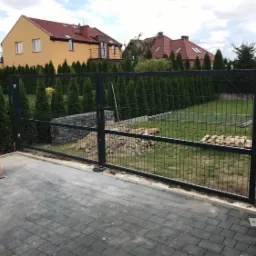 Nowo zamontowana, ciemnoszara, dwuskrzydłowa brama z paneli ogrodzeniowych na posesji z kostki brukowej, widoczne rury PCV i częściowo ułożony murek z bloczków betonowych w tle.