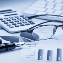 Biurko z kalkulatorem, okularami, piórem wiecznym i wykresami słupkowymi oraz kołowymi w odcieniach niebieskiego.