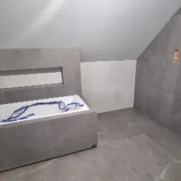 Surowa łazienka w trakcie wykańczania, szare płytki imitujące marmur na ścianach i podłodze, biała wanna zabezpieczona folią, widoczne punkty instalacji wodnej, wnęka w zabudowie wanny.