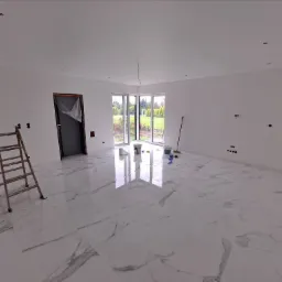 Wnętrze pokoju po malowaniu ścian na biało, widoczne marmurowe płytki na podłodze, drabina, okno z widokiem na ogród, puszki z farbą i akcesoria malarskie.