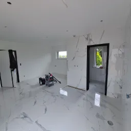 Wnętrze w trakcie prac wykończeniowych: białe płytki imitujące marmur na podłodze i ścianach, widoczny odkurzacz przemysłowy i narzędzia, czarne futryny drzwiowe, surowe gniazdka elektryczne...