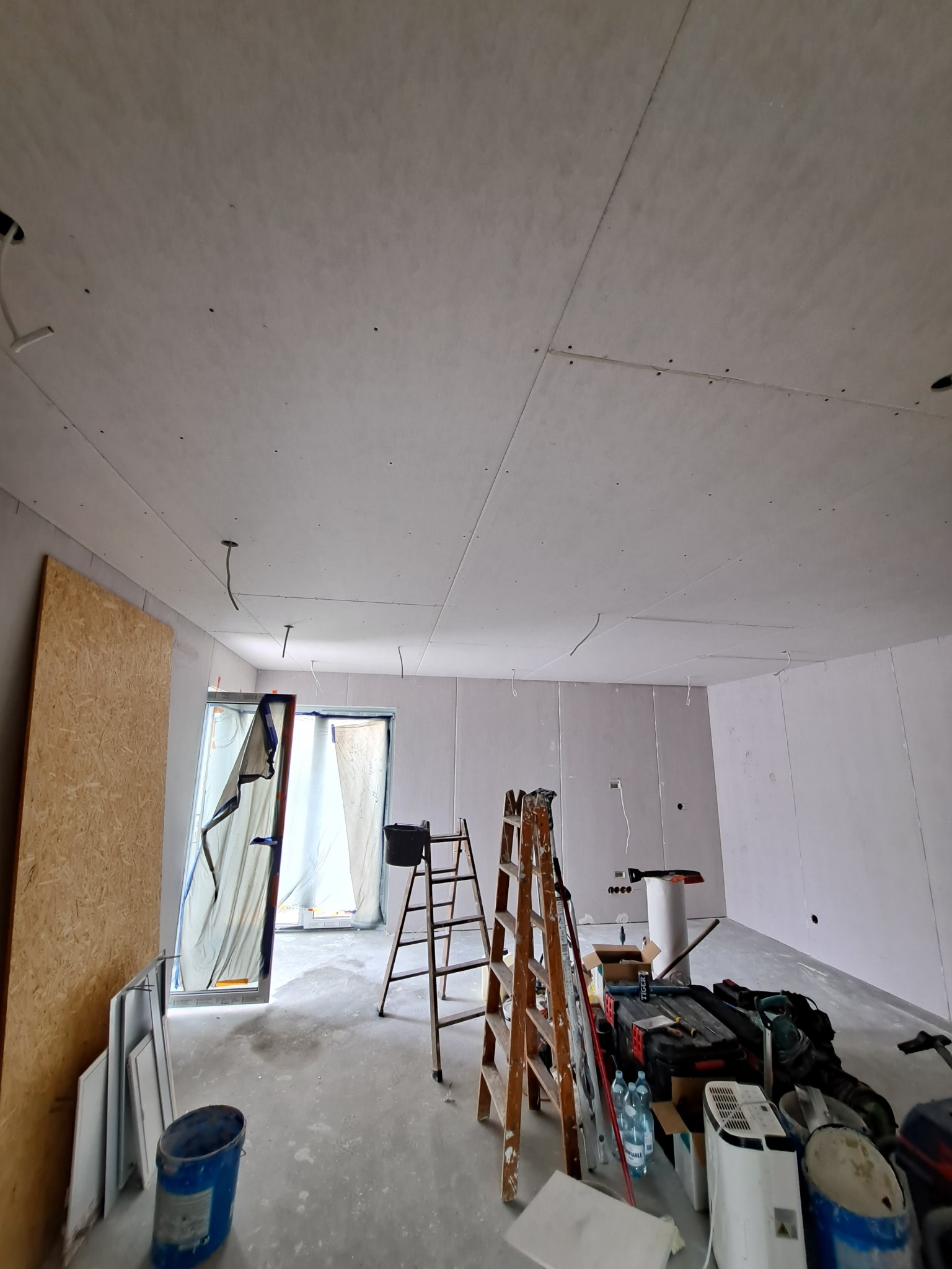 Pokój w trakcie prac wykończeniowych: zamontowane płyty gipsowo-kartonowe na suficie, widoczne drabiny, narzędzia i materiały budowlane, okno zabezpieczone folią.