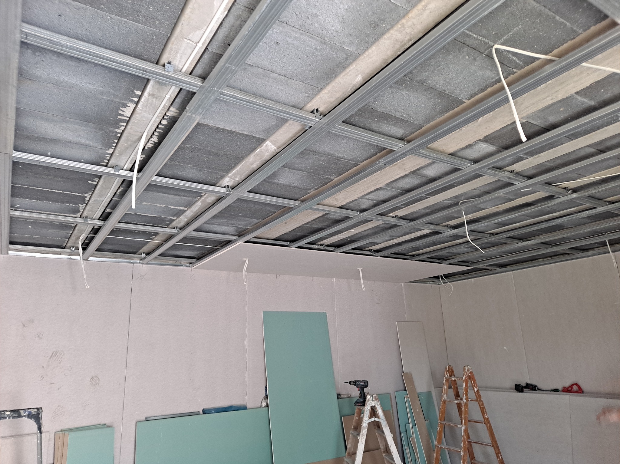 Montaż sufitu podwieszanego z płyt gipsowo-kartonowych na metalowej konstrukcji, widoczne płyty, wiertarka na drabinie i kable zwisające z sufitu.