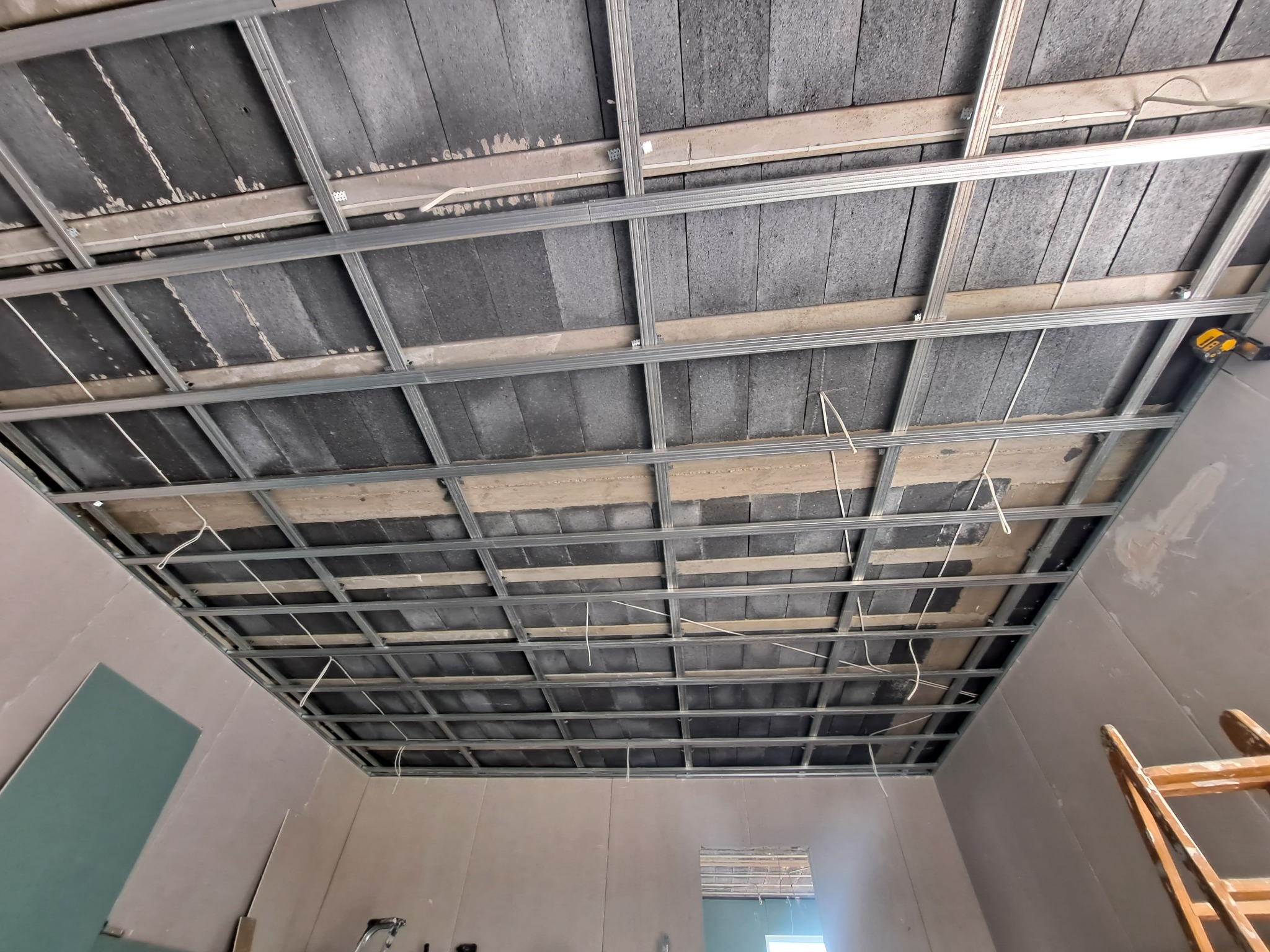 Sufit podwieszany w trakcie montażu: widoczne metalowe profile, izolacja akustyczna i częściowo zamontowane płyty gipsowo-kartonowe, z wiszącymi przewodami elektrycznymi.