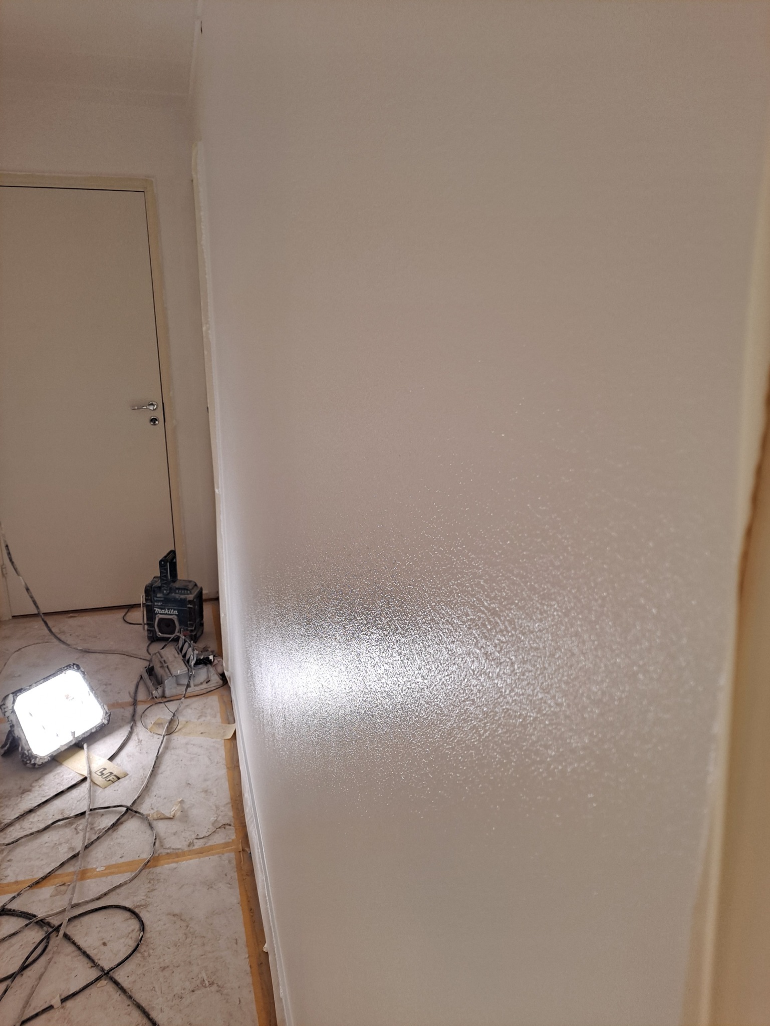 Świeżo pomalowana, biała ściana w trakcie schnięcia, oświetlona lampą budowlaną Makita, widoczne przewody i zabezpieczona podłoga.