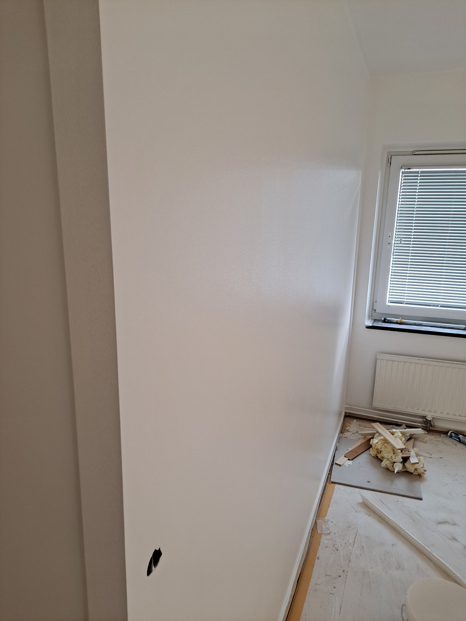 Świeżo pomalowana na biało ściana w pokoju z widocznym gniazdkiem elektrycznym, okno z żaluzjami i grzejnik. Na podłodze pozostałości po pracach wykończeniowych: fragmenty listew przypodłogowych...