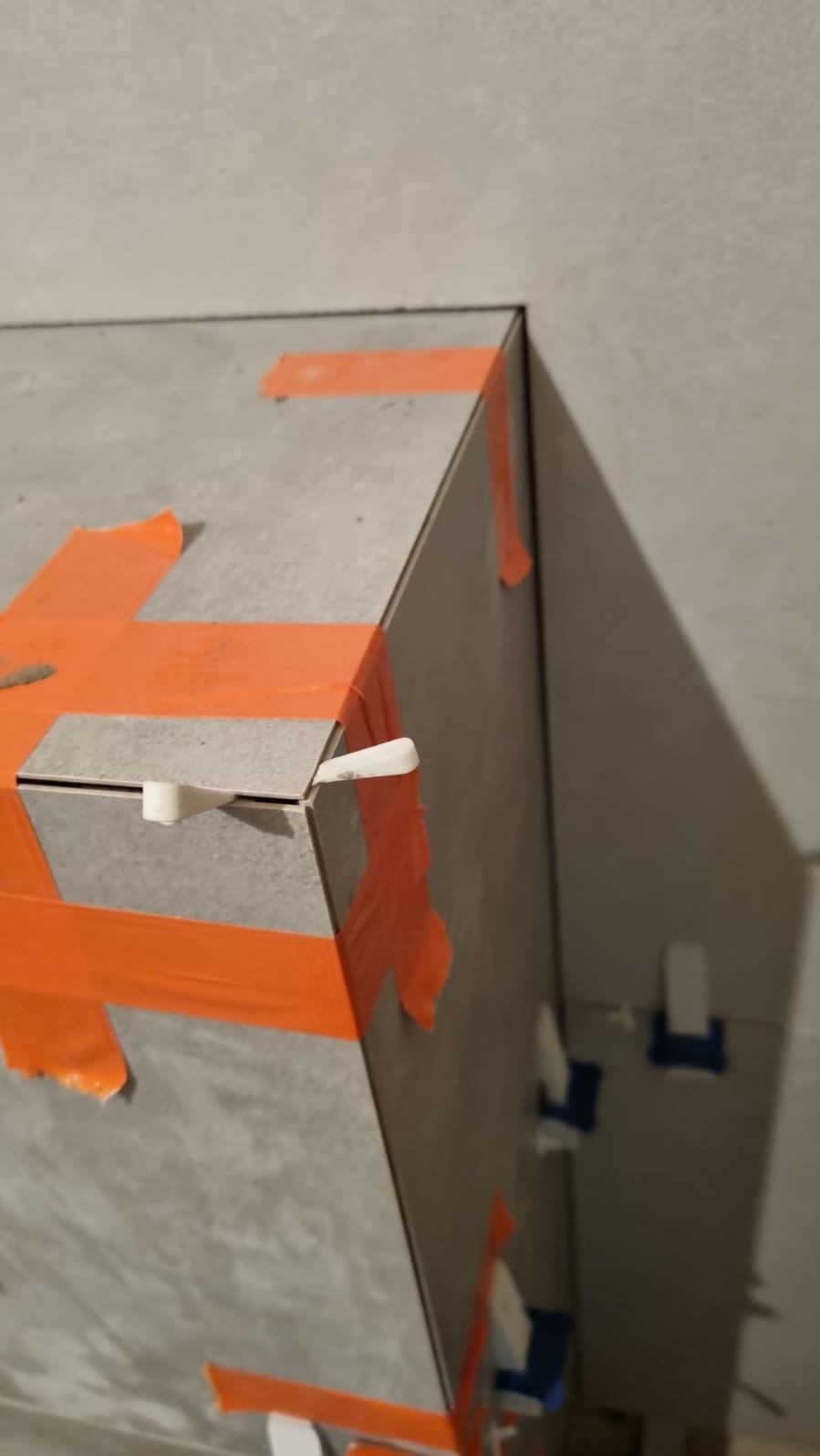 Szare płytki ceramiczne na obudowie w trakcie montażu, zabezpieczone pomarańczową taśmą i białymi krzyżykami dystansowymi.