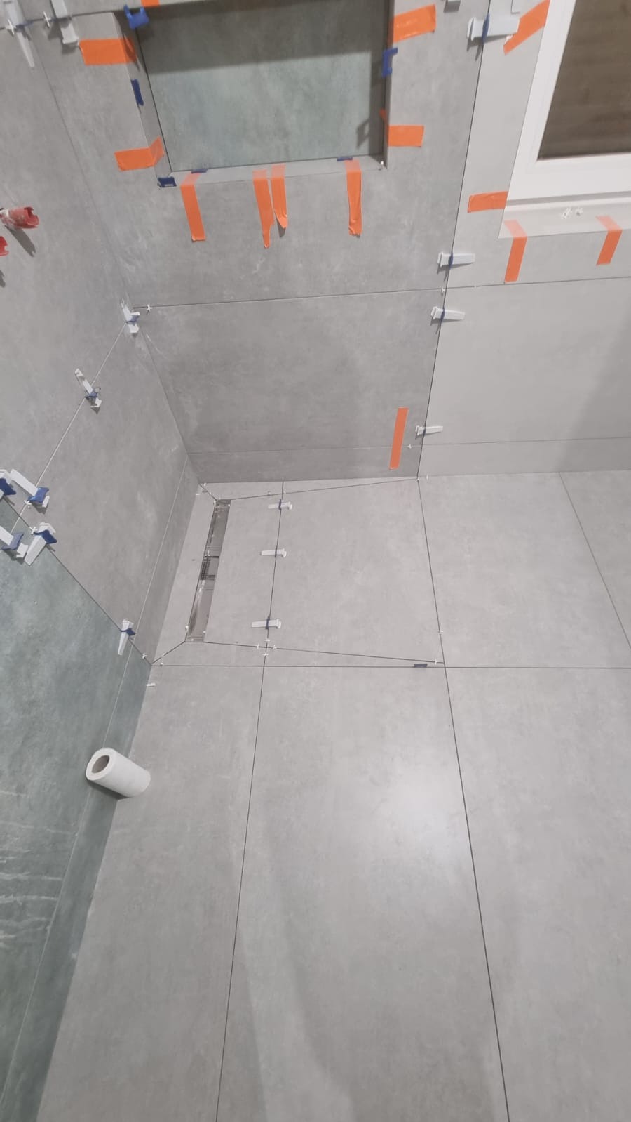 Szare płytki wielkoformatowe w trakcie układania w łazience, widoczne systemy poziomowania płytek (klipsy i kliny) oraz taśma malarska zabezpieczająca narożniki, odpływ liniowy w podłodze.