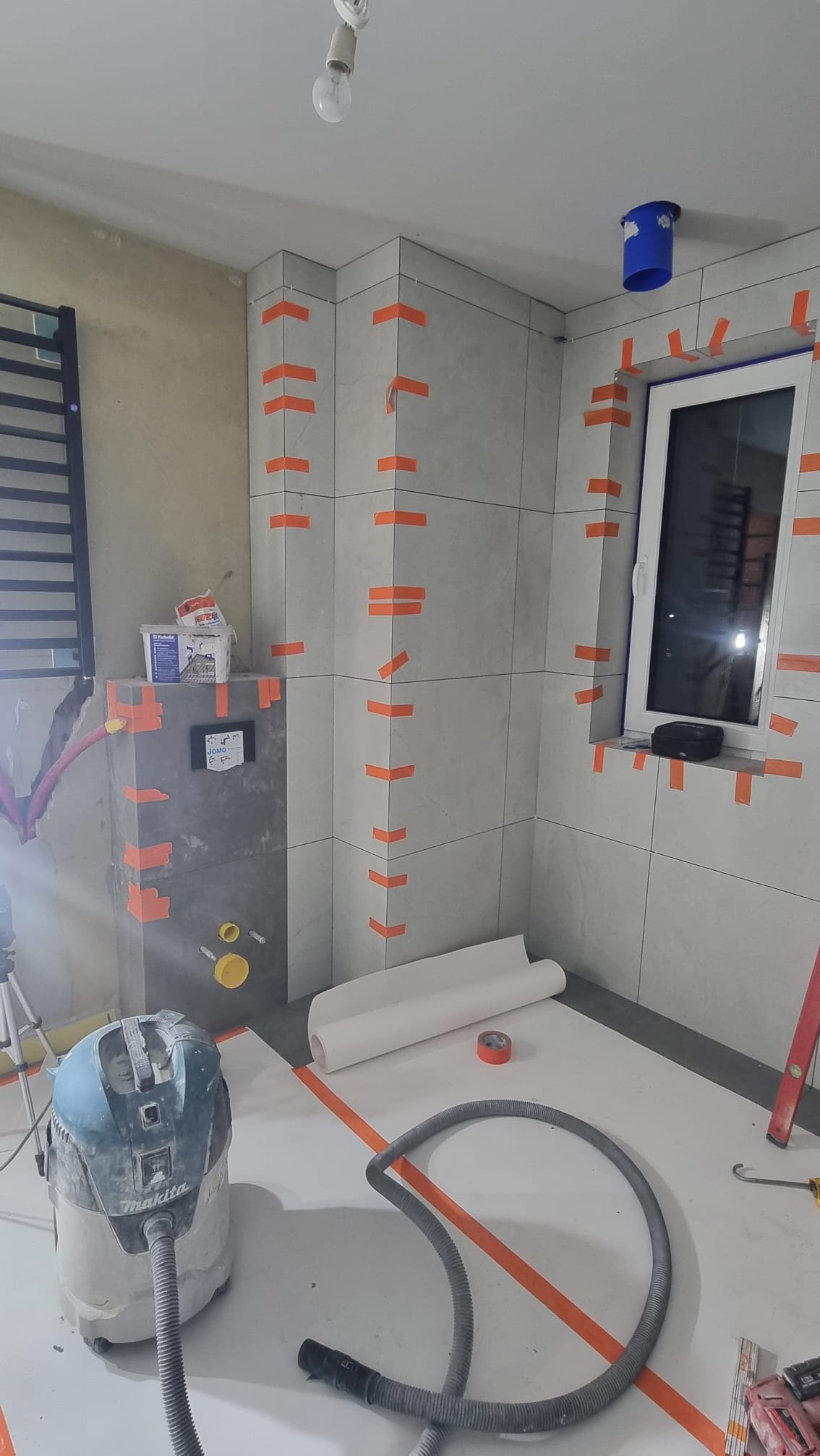 Łazienka w trakcie remontu: szare płytki na ścianach, zabezpieczone pomarańczową taśmą, widoczny stelaż podtynkowy WC, odkurzacz przemysłowy Makita i rolka papieru zabezpieczającego podłogę.