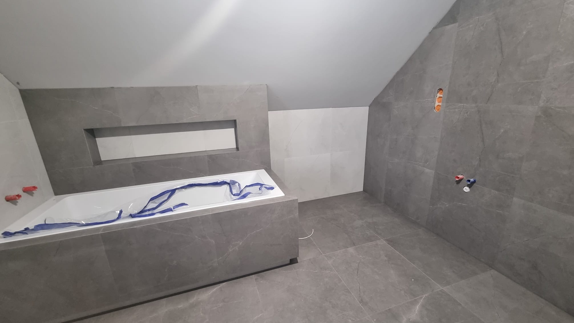 Surowa łazienka w trakcie wykańczania, szare płytki imitujące marmur na ścianach i podłodze, biała wanna zabezpieczona folią, widoczne punkty instalacji wodnej, wnęka w zabudowie wanny.