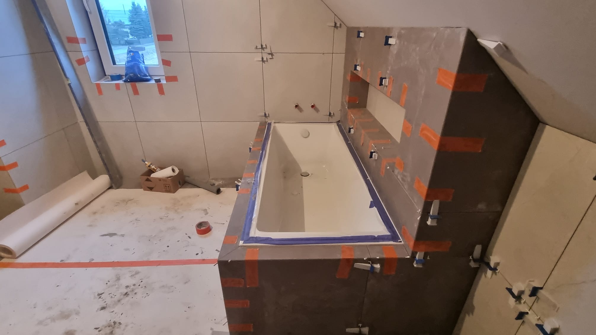 Ukośna łazienka w trakcie układania płytek: widoczna wanna z niebieską taśmą zabezpieczającą, ściany wyłożone jasnymi płytkami z pomarańczową taśmą, poziomica oparta o ścianę, karton z akcesoriami...