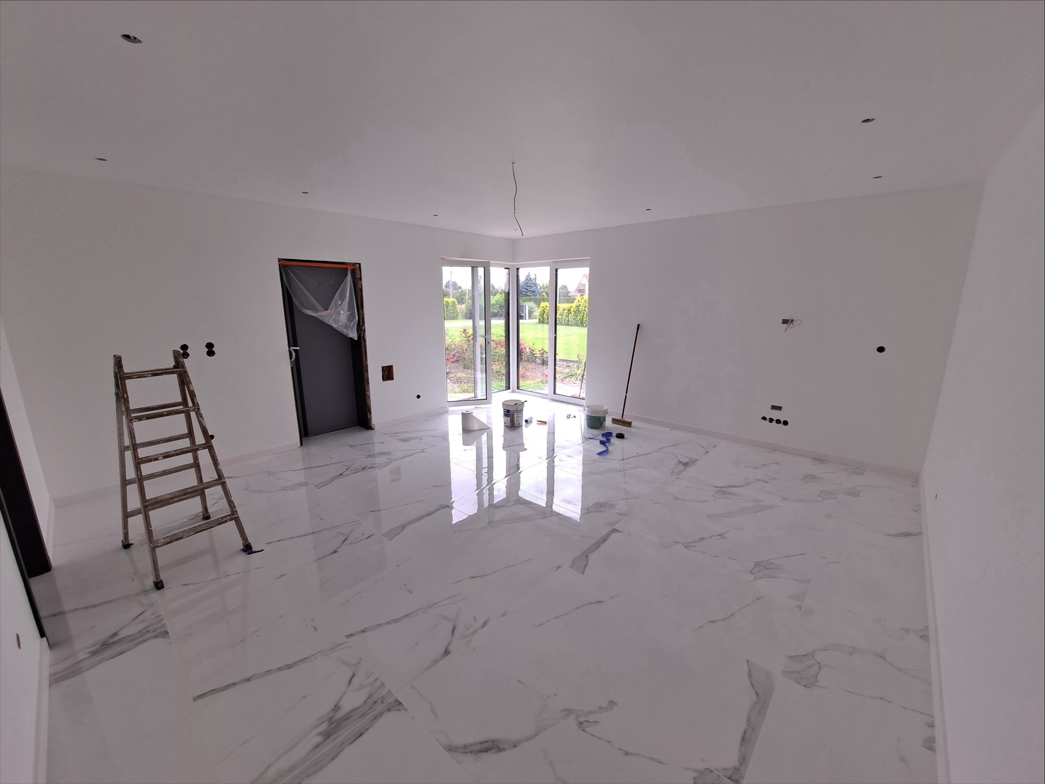 Wnętrze pokoju po malowaniu ścian na biało, widoczne marmurowe płytki na podłodze, drabina, okno z widokiem na ogród, puszki z farbą i akcesoria malarskie.