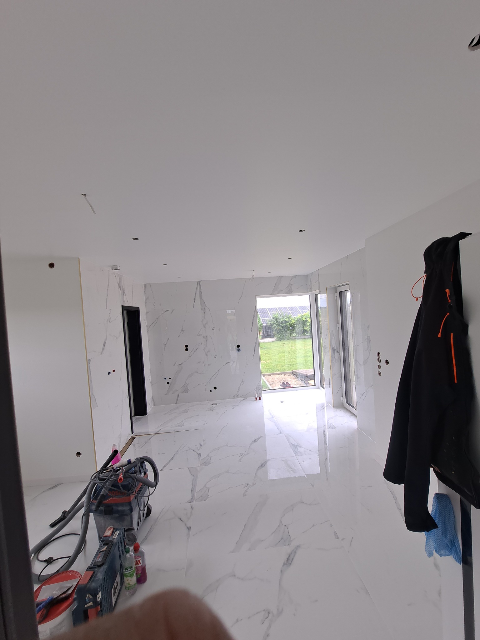 Wnętrze w trakcie prac wykończeniowych: białe marmurowe płytki na podłodze i ścianach, widoczne narzędzia budowlane, odkurzacz przemysłowy i płyny czyszczące, czarna kurtka wisząca na ścianie, okno...