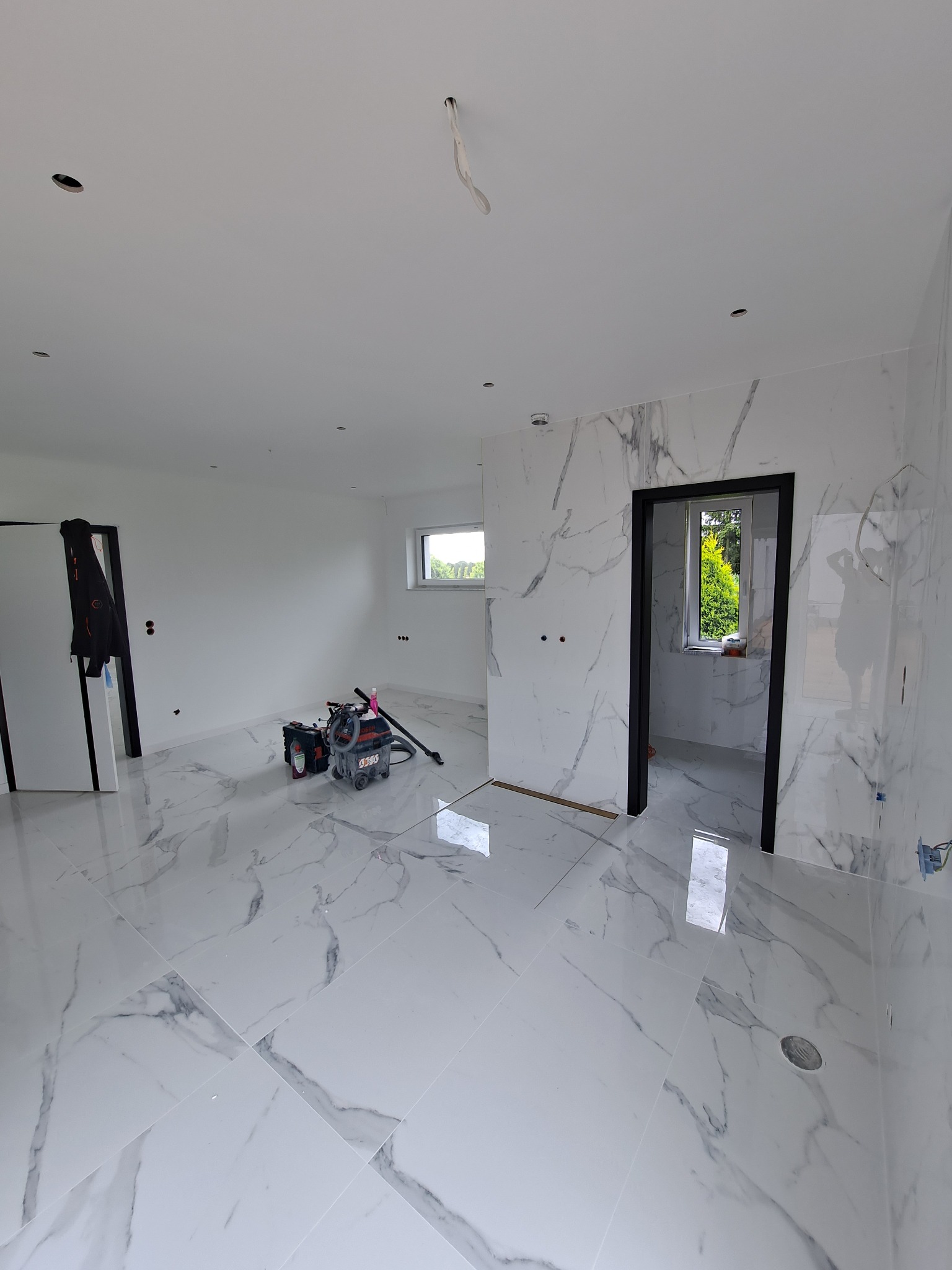 Wnętrze w trakcie prac wykończeniowych: białe płytki imitujące marmur na podłodze i ścianach, widoczny odkurzacz przemysłowy i narzędzia, czarne futryny drzwiowe, surowe gniazdka elektryczne...