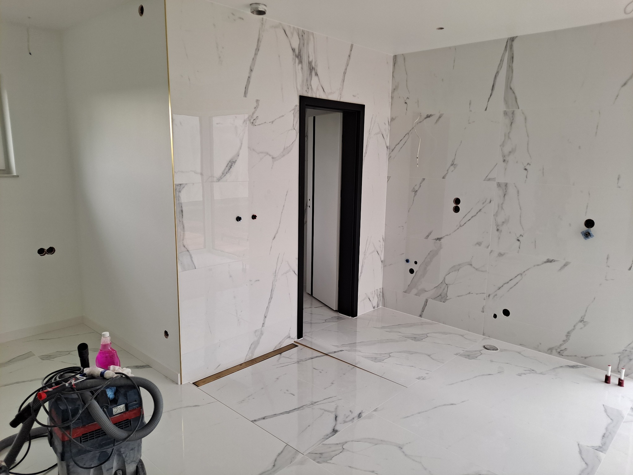 Wnętrze łazienki w trakcie prac wykończeniowych: białe marmurowe płytki na ścianach i podłodze, czarna futryna drzwiowa, widoczny odkurzacz przemysłowy i butelka różowego płynu.