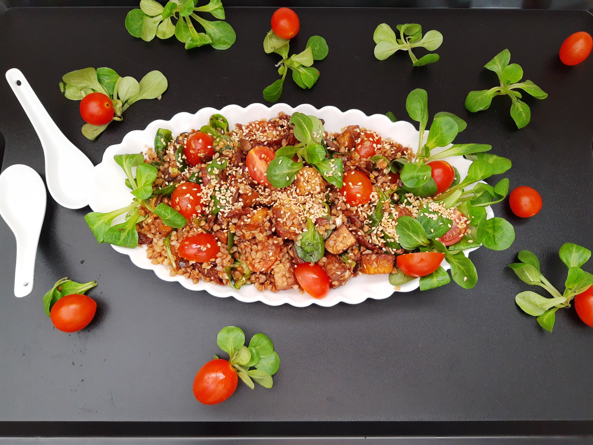 Sałatka z kaszy gryczanej, tofu, pomidorków cherry i roszponki, posypana sezamem, podana na białym talerzu z falbanką, obok dwie białe łyżki ceramiczne, na czarnym tle.