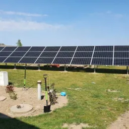 Długi rząd paneli słonecznych na metalowej konstrukcji, zainstalowany na trawiastym terenie z widocznymi narzędziami ogrodniczymi i elementami infrastruktury podziemnej.