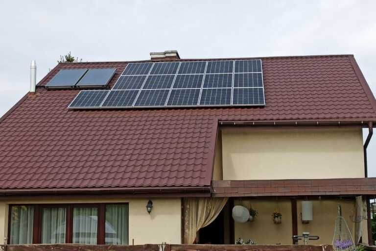 Panele słoneczne zamontowane na brązowym dachu domu jednorodzinnego z beżową elewacją, widoczne okna i fragment balkonu.