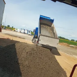 Ciężarówka z otwartą skrzynią wyładowuje ziemię na placu budowy, w tle widoczne baraki i pole uprawne.