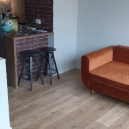 Jasne panele podłogowe ułożone w salonie z ceglanym elementem dekoracyjnym i pomarańczową sofą, widoczne dwa czarne hokery przy blacie.