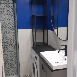 Nowoczesna łazienka z niebieską ścianą, umywalką nablatową na szafce w kolorze szarym, pralką pod blatem, czarnym regałem i kabiną prysznicową z wzorzystą szybą, wykończona płytkami podłogowymi...