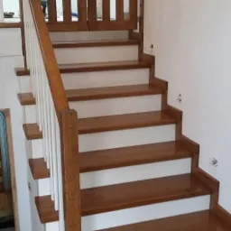 Drewniane schody z barierką i zabezpieczającą bramką na szczycie, stopnie w kolorze brązowym z białymi podstopnicami, białe słupki balustrady.