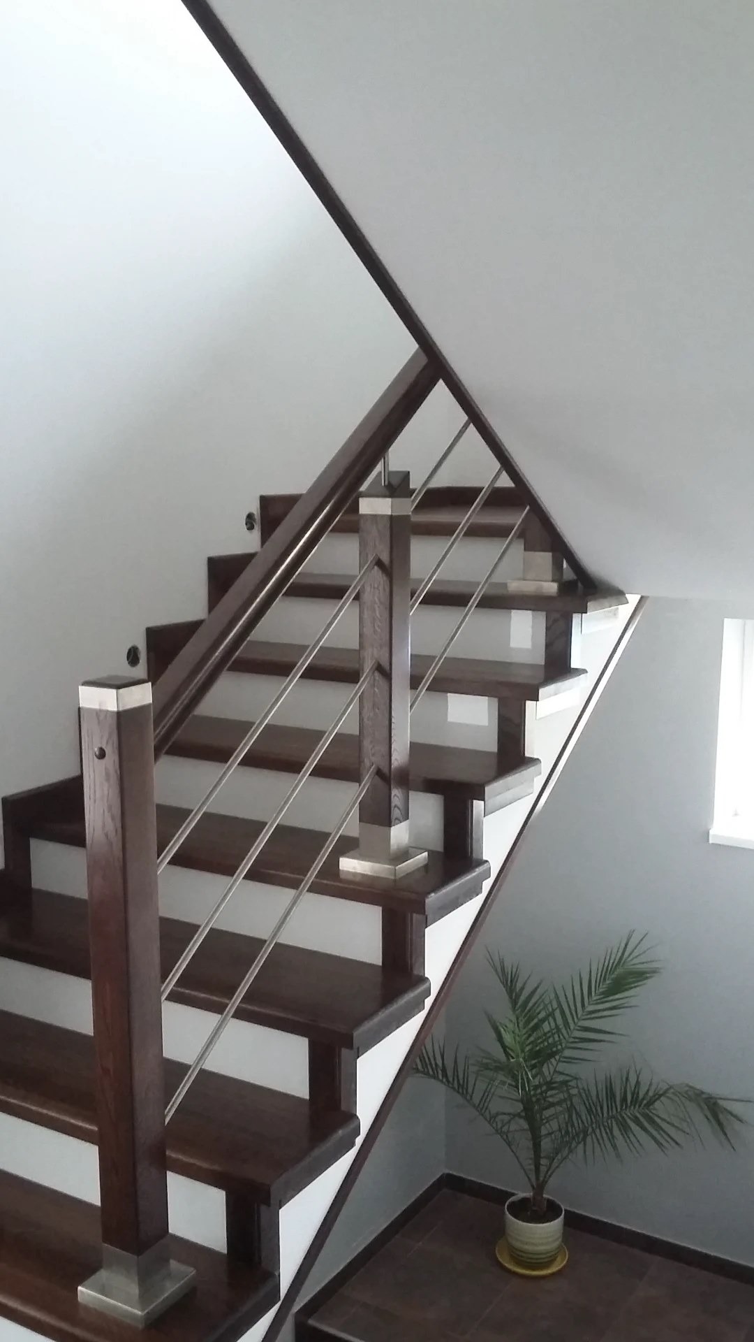 Nowoczesne schody z ciemnego drewna z metalowymi relingami i dekoracyjnymi słupkami, oświetlone naturalnym światłem w jasnym wnętrzu z rośliną doniczkową u podnóża.