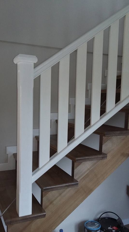 Widok z dołu na fragment schodów z drewnianymi stopniami w odcieniach brązu i jasnego drewna, z białymi balustradami i poręczą, przy szarej ścianie, z widocznymi puszkami farby na dole.