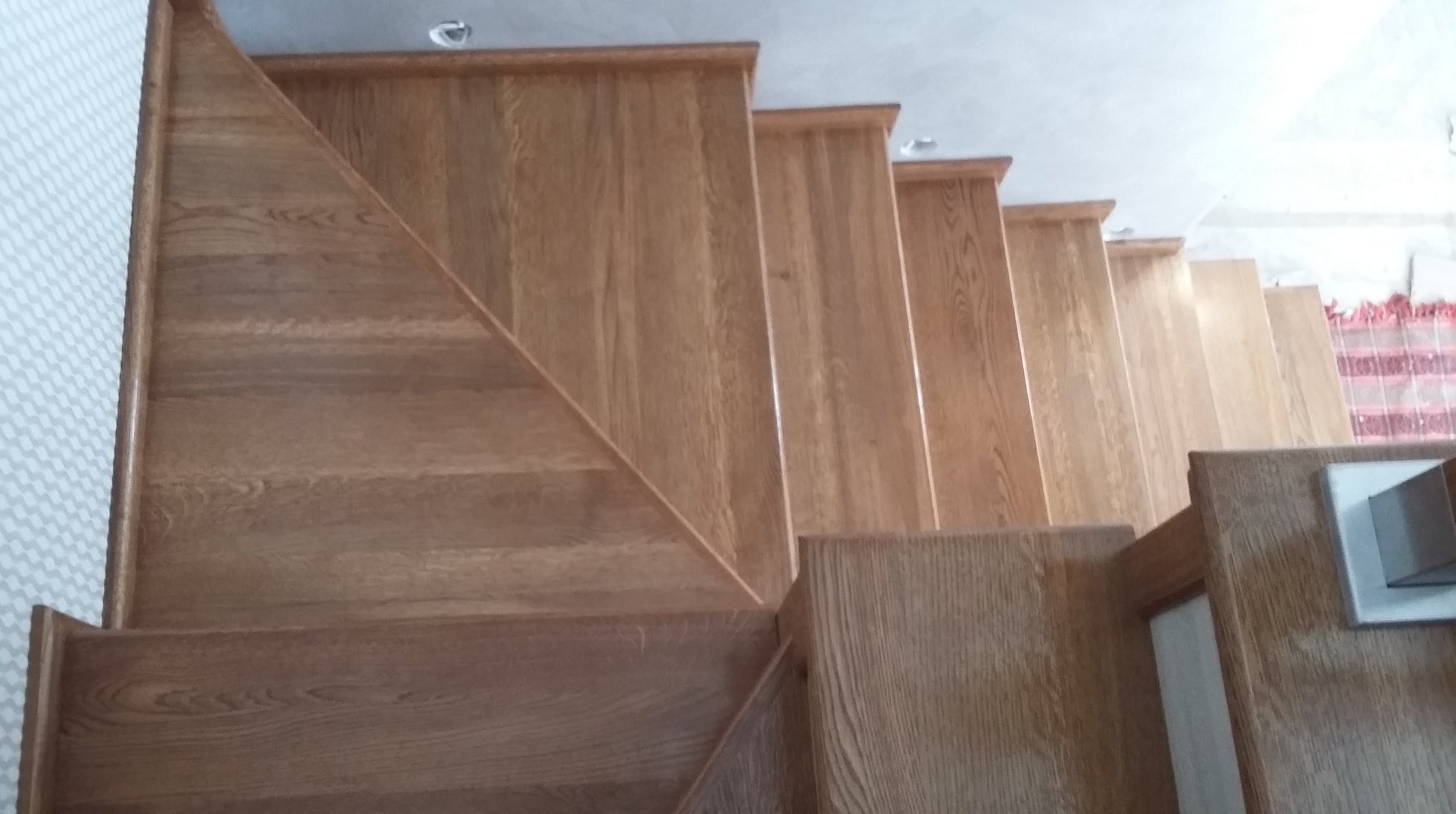 Widok z góry na jasne, dębowe schody z drewnianą barierką, z widoczną fakturą drewna i subtelnym oświetleniem sufitowym.