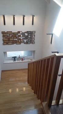 Jasne, drewniane stopnie schodów z balustradą, prowadzące w górę, oświetlone przez lampy ścienne w kształcie wsporników. Na ścianie dekoracja z drewnianych elementów. Na parapecie okna widoczne...