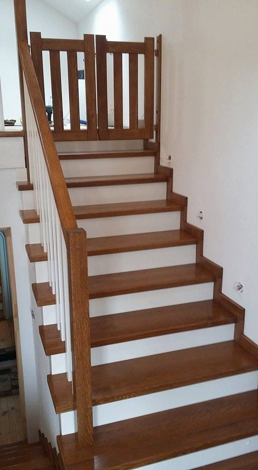 Drewniane schody z barierką i zabezpieczającą bramką na szczycie, stopnie w kolorze brązowym z białymi podstopnicami, białe słupki balustrady.